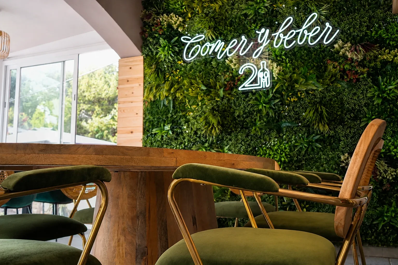 Rincón terminado del restaurante Los Dos en El Campello con jardín vertical de plantas, neón «Comer y beber», mesa de madera y sillas tapizadas en verde
